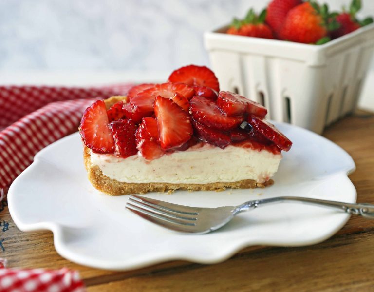 A slice of strawberry-topped cheesecake on a white plate with a fork, with a container of strawberries and a red checkered cloth in the background.
