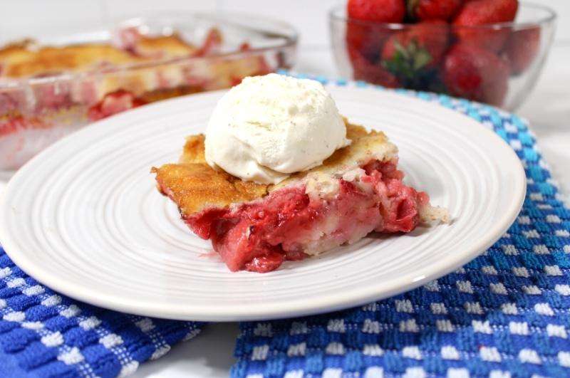 A slice of strawberry cobbler topped with a scoop of vanilla ice cream on a white plate, with fresh strawberries and more cobbler in the background.