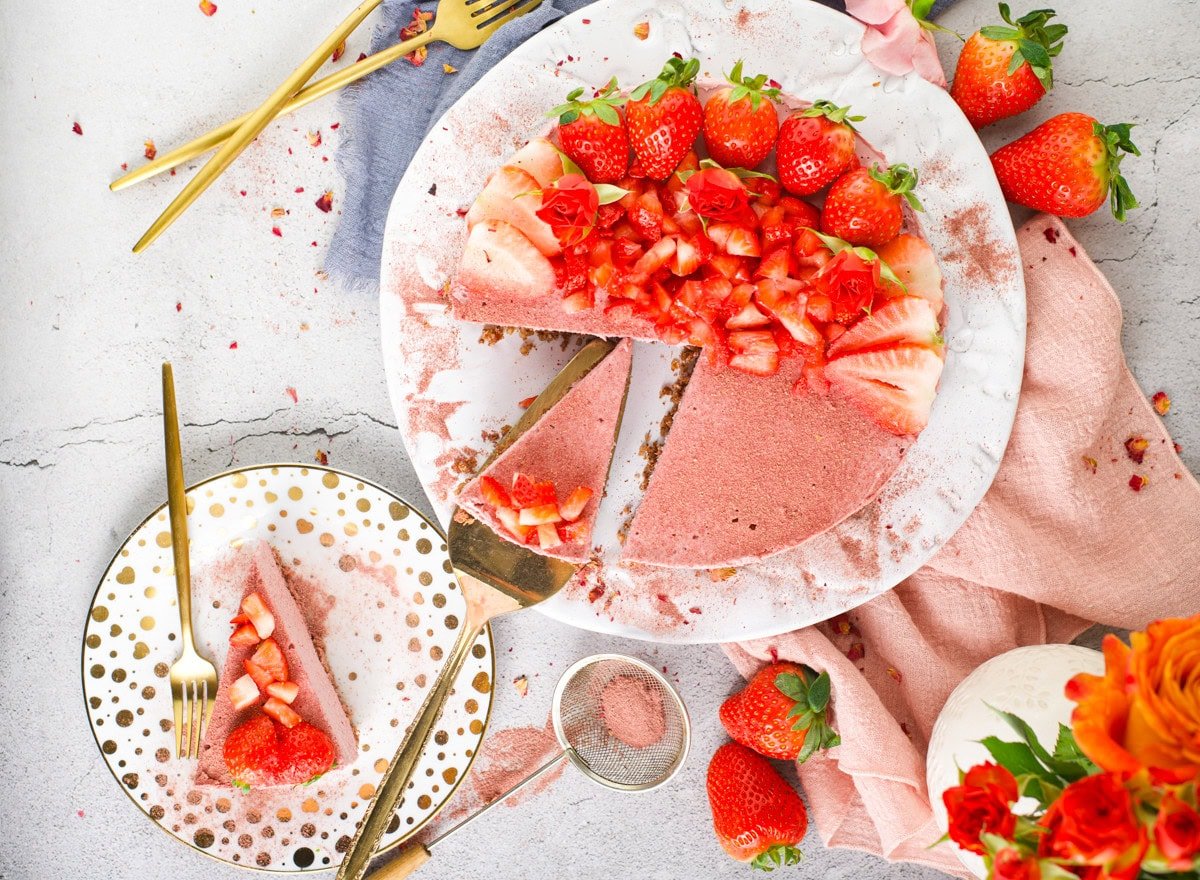 A round strawberry cake with a slice removed sits on a plate, topped with fresh strawberries. A slice is served on a polka dot plate, surrounded by forks and scattered strawberries.