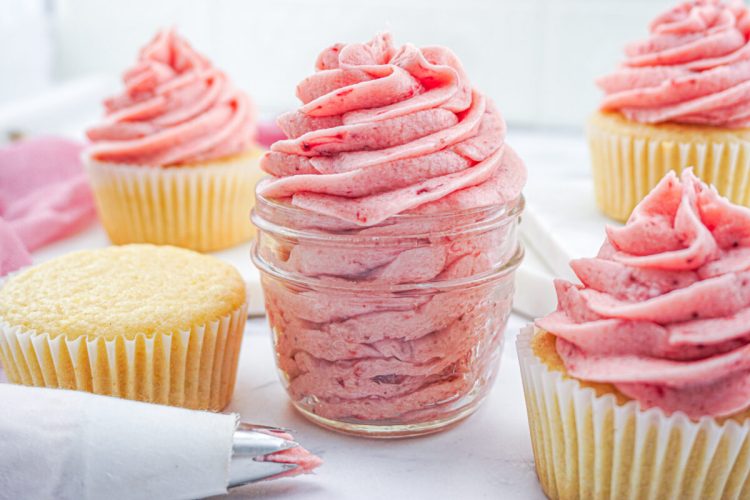 A small glass jar filled with pink frosting is surrounded by vanilla cupcakes, some with and some without pink frosting. A piping bag with pink frosting lies nearby.