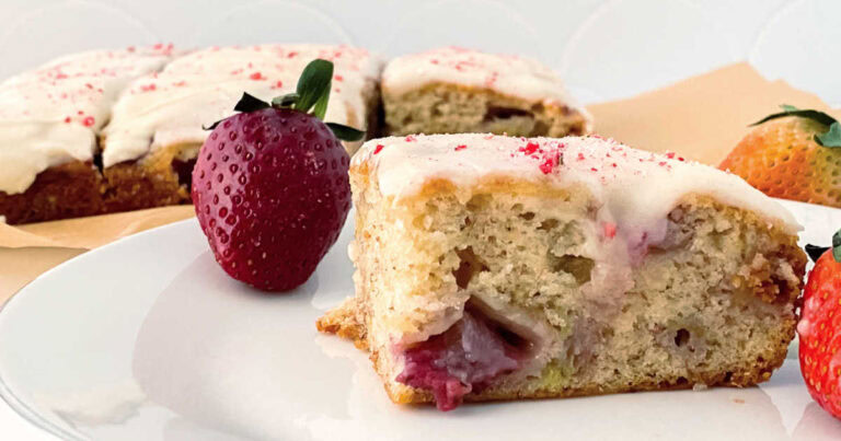 A slice of strawberry cake with white icing sits on a plate next to fresh strawberries, with more iced cake pieces in the background.