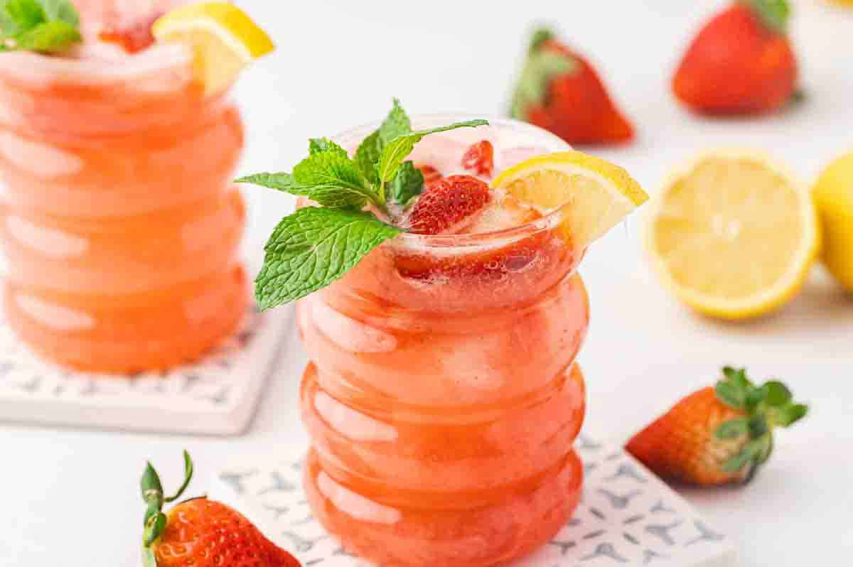 A glass of pink strawberry lemonade garnished with a lemon slice, mint leaves, and strawberry, with whole strawberries and lemons in the background.