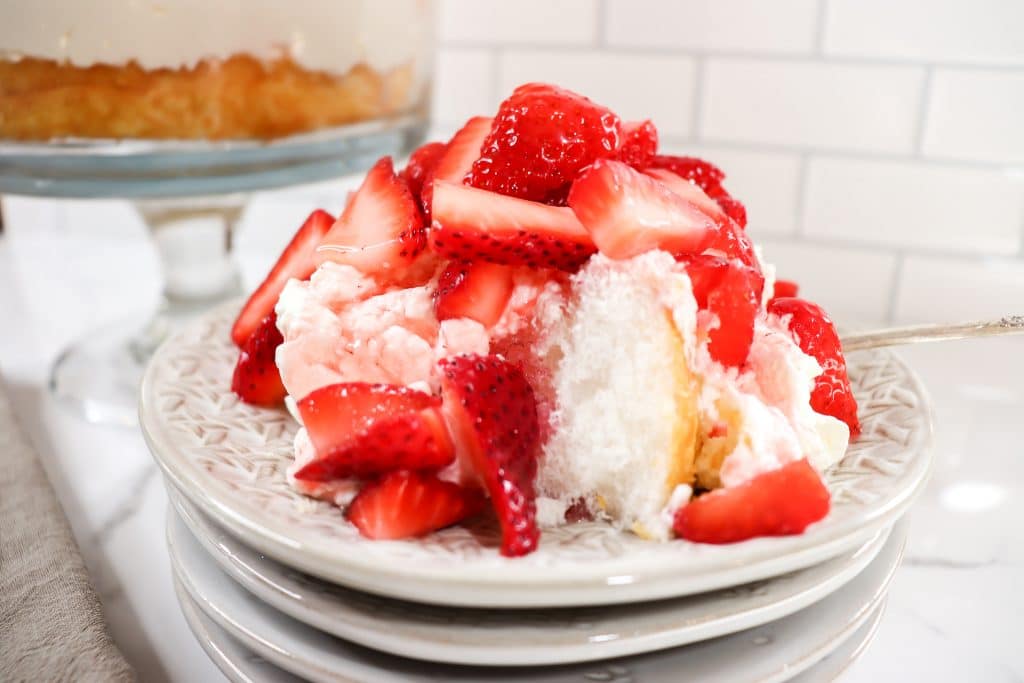 A slice of strawberry shortcake with whipped cream on a plate, topped with fresh strawberries, next to a larger cake in the background.