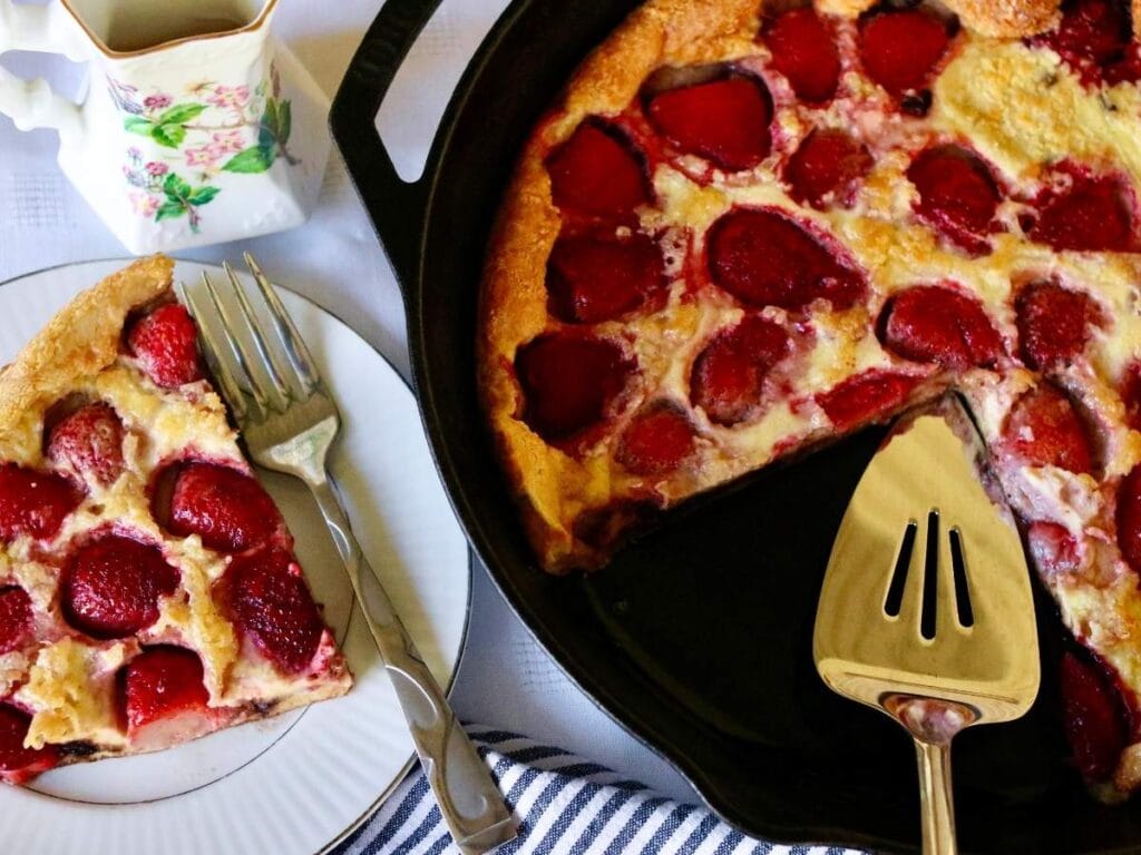 A slice of strawberry clafoutis on a plate with a fork, next to a cast iron skillet containing the rest of the dessert and a pie server. A small floral pitcher is also visible.
