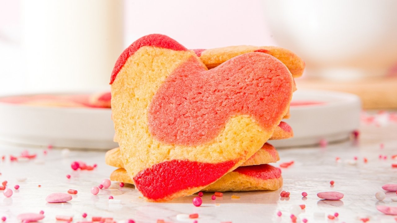 A stack of heart-shaped cookies with red and yellow marbled patterns, surrounded by pink and red sprinkles on a white surface.