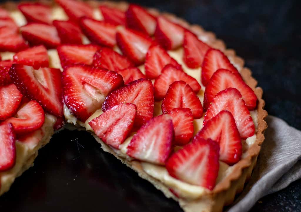 A strawberry tart with a slice missing, featuring sliced strawberries arranged on top of a creamy filling in a pastry crust.