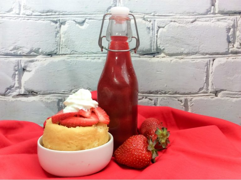 A dessert with whipped cream and sliced strawberries sits in a bowl next to a bottle of strawberry sauce and two whole strawberries on a red cloth, against a white brick wall.