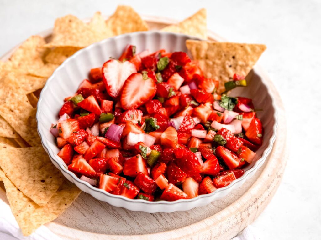 A bowl of strawberry salsa with chopped strawberries, onions, and herbs, served with tortilla chips on a round wooden board.