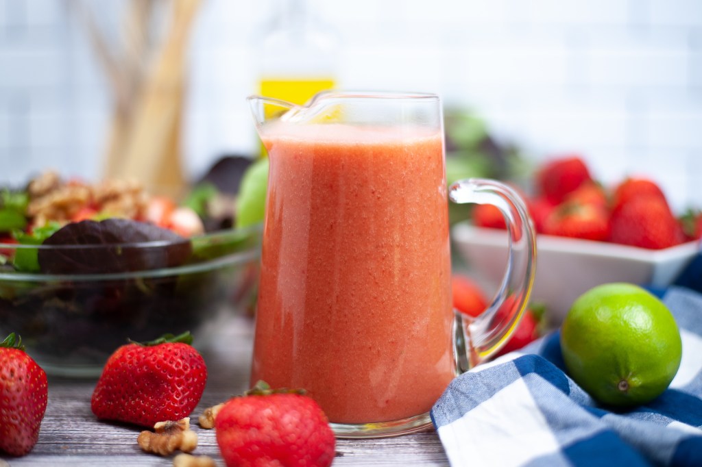 A glass pitcher filled with strawberry smoothie sits on a table surrounded by fresh strawberries, a lime, walnuts, and a salad bowl, with a blue and white checkered cloth nearby.