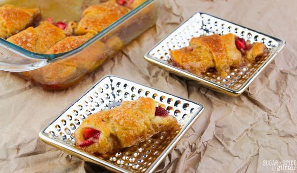 Two small square plates each hold a strawberry-filled pastry, with a glass baking dish containing more pastries in the background on a crumpled paper surface.