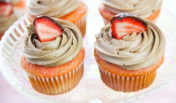 Two pink cupcakes with swirled brown frosting, each topped with a sliced strawberry, displayed on a glass plate.