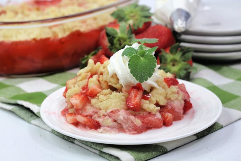 A serving of strawberry crumble topped with whipped cream and a mint leaf sits on a white plate with fresh strawberries in the background.