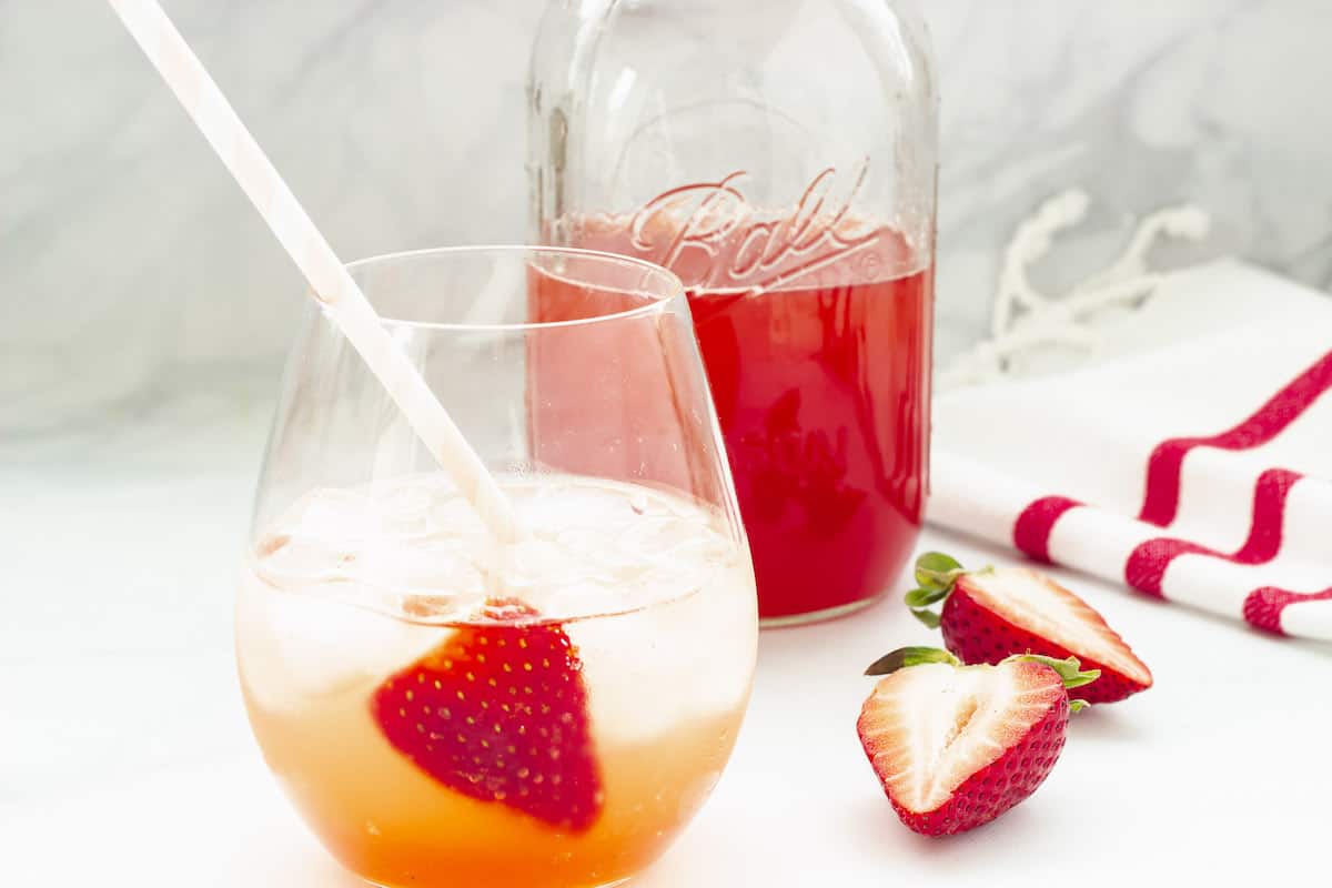 A glass of iced strawberry drink with a straw and a strawberry slice, next to a jar of red liquid and two halved strawberries on a white surface.
