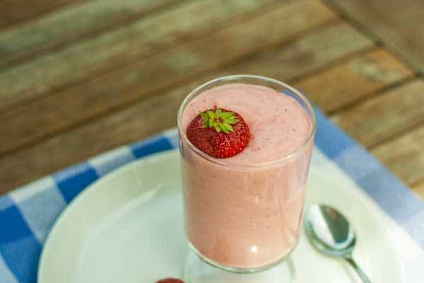 A glass of strawberry mousse topped with a whole strawberry, placed on a white plate with a spoon on the side.