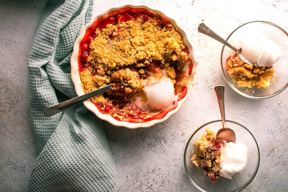 A baked fruit crumble in a round dish with two servings in bowls, each topped with a scoop of vanilla ice cream, next to a light green kitchen towel.