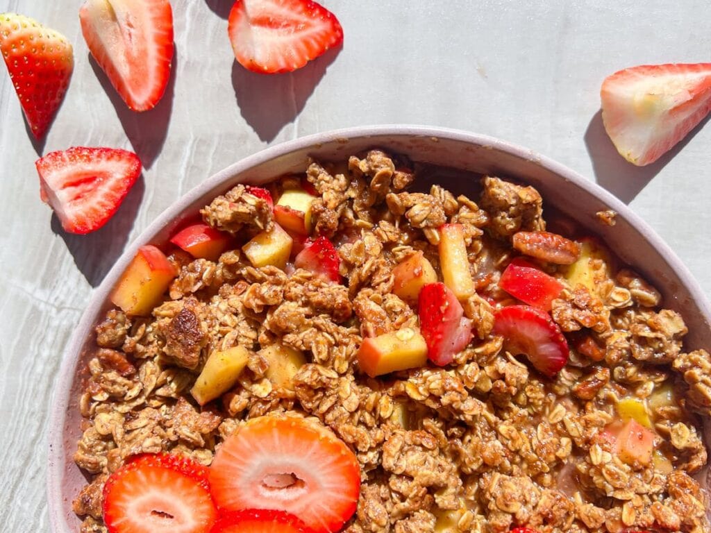A close-up of a bowl of granola with sliced strawberries and pieces of fruit, with additional strawberry halves scattered on the surface nearby.