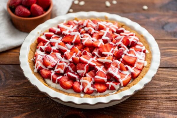A strawberry pie topped with sliced strawberries and white icing drizzle, in a white pie dish on a wooden table. A bowl of strawberries is in the background.