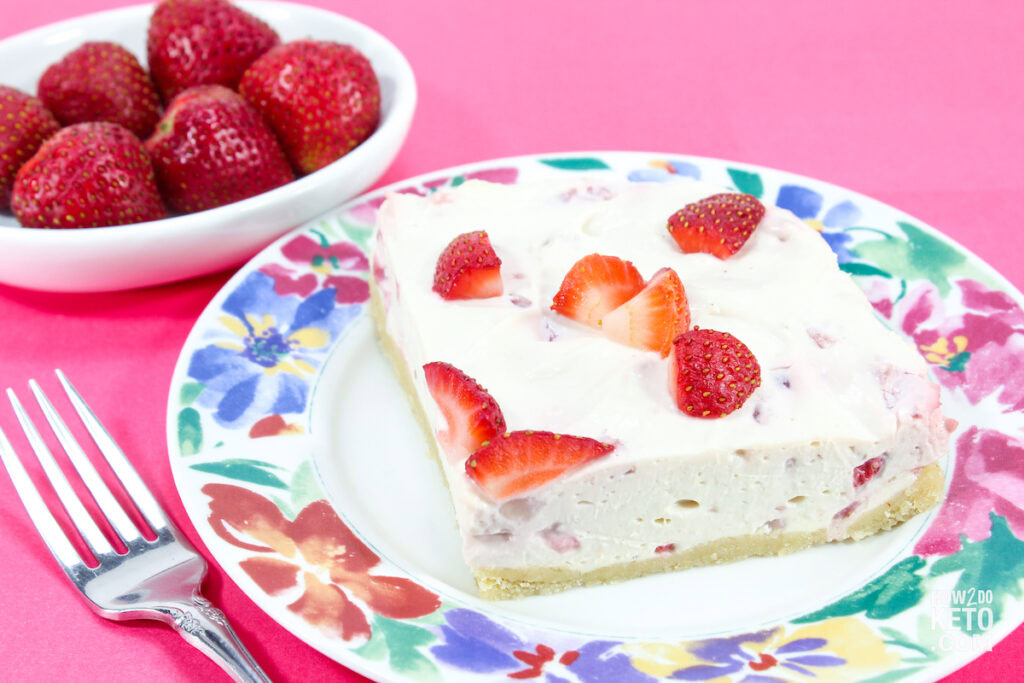 A slice of strawberry cheesecake topped with fresh strawberry pieces on a floral plate, with a bowl of whole strawberries and a fork beside it on a pink background.