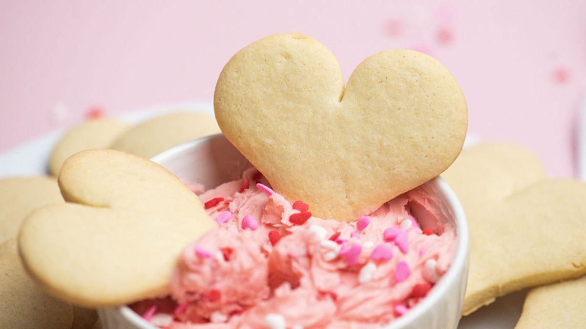 Heart-shaped sugar cookies with pink frosting and sprinkles in a white bowl, surrounded by more cookies, against a pink background.