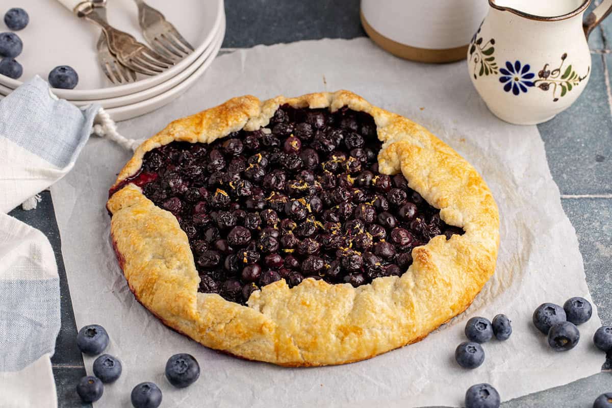A rustic blueberry galette with a golden, folded crust sits on parchment paper, surrounded by fresh blueberries, plates, forks, and a decorative pitcher.