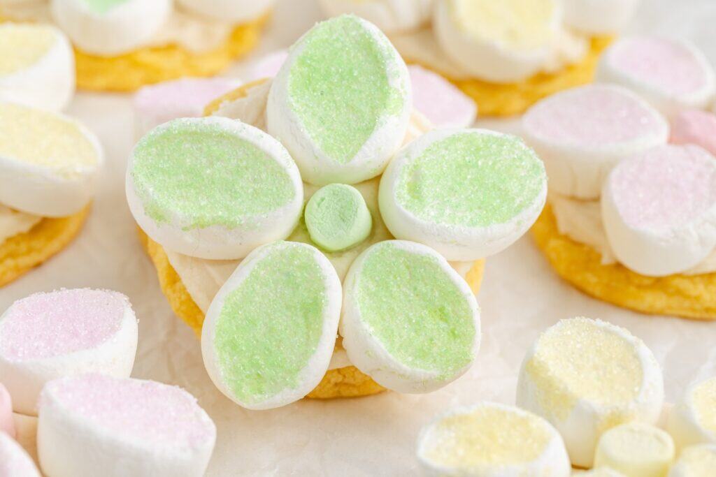 A cookie decorated with marshmallows arranged in the shape of a flower, featuring green and white petals and a green center, with other cookies and marshmallows in the background.