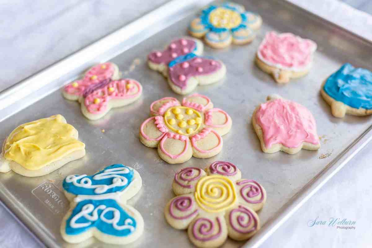 A baking tray with ten decorated sugar cookies in shapes like flowers, butterflies, and animals, each topped with colorful icing designs.