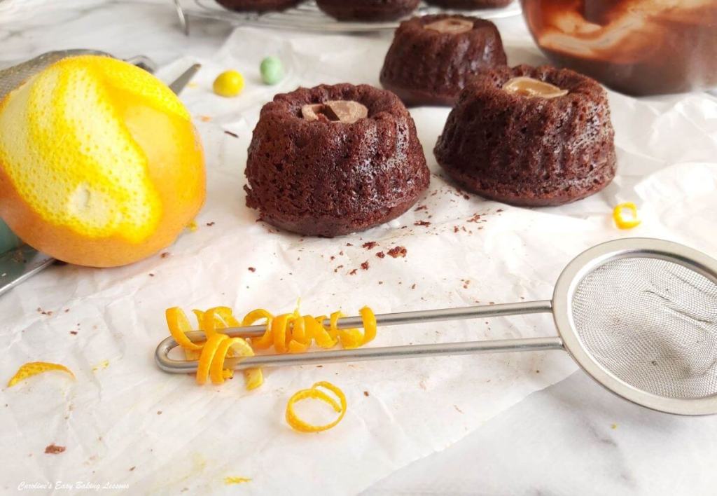 Two mini chocolate bundt cakes sit on parchment paper next to a zested lemon, a metal strainer, and strips of lemon zest.