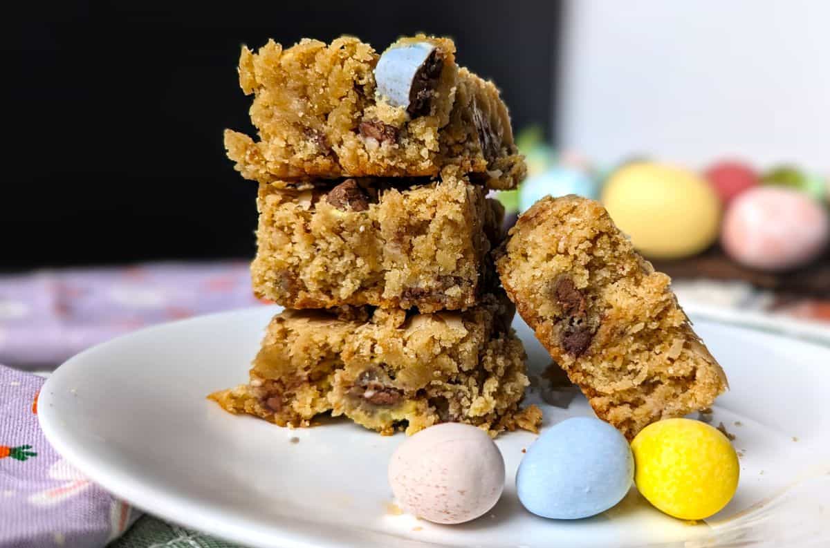 A stack of four cookie bars with chocolate pieces on a white plate, accompanied by three pastel-colored candy eggs.