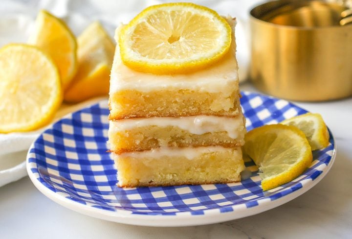 A stack of three lemon bars with icing, topped with a lemon slice, sits on a blue and white checkered plate with lemon wedges in the background.