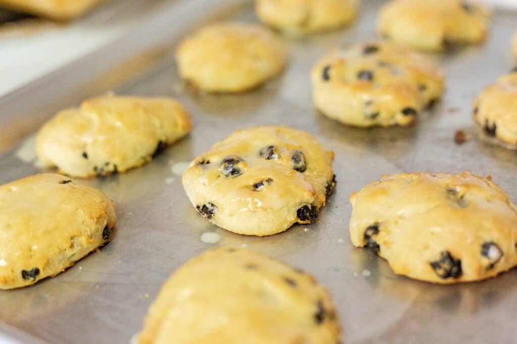 A baking tray with freshly baked, golden-brown scones containing dark dried fruit, possibly currants or raisins, with a shiny glaze on top.