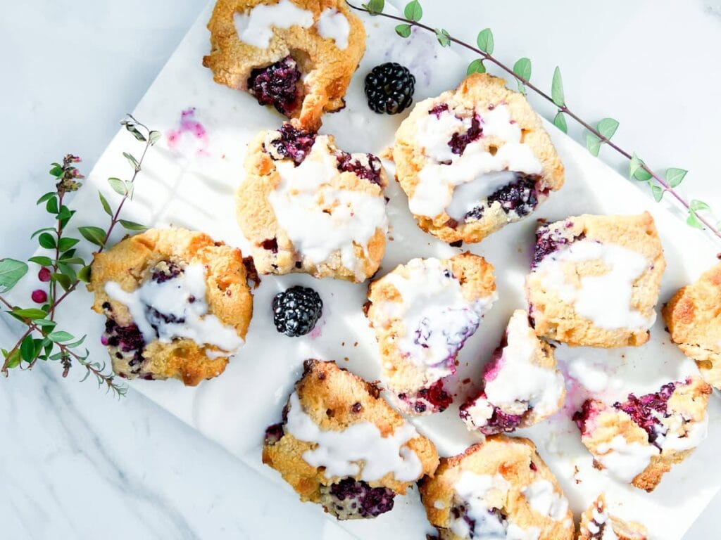 A marble tray with blackberry scones topped with white icing, fresh blackberries, and a sprig of greenery on a white surface.