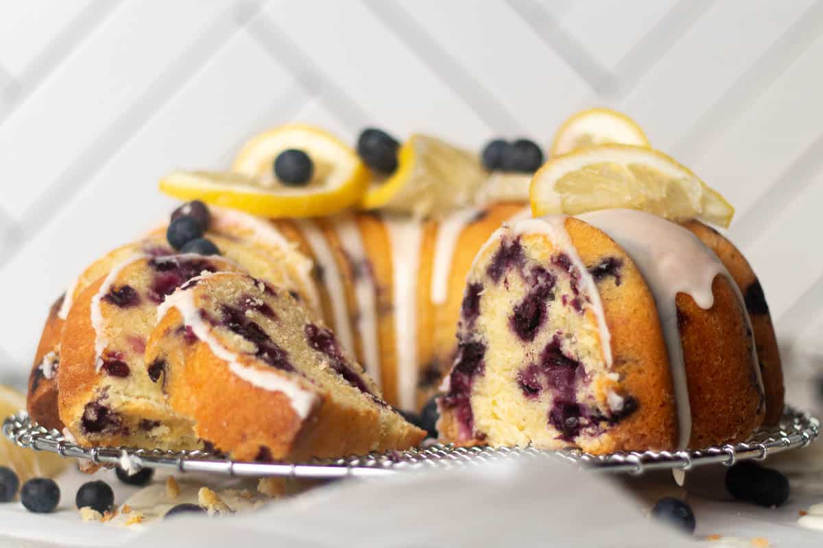 Bundt cake with blueberries and icing, garnished with lemon slices and fresh blueberries, displayed on a wire rack.