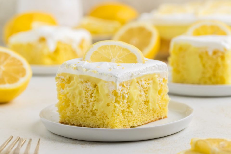 A slice of lemon cake with creamy filling and whipped topping, garnished with a lemon wheel, sits on a white plate.