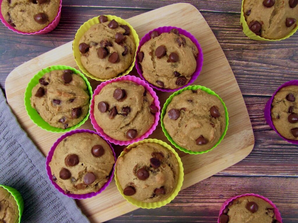 Nine chocolate chip muffins in colorful silicone baking cups are arranged on a wooden cutting board, with a few more muffins around it on a wooden surface.