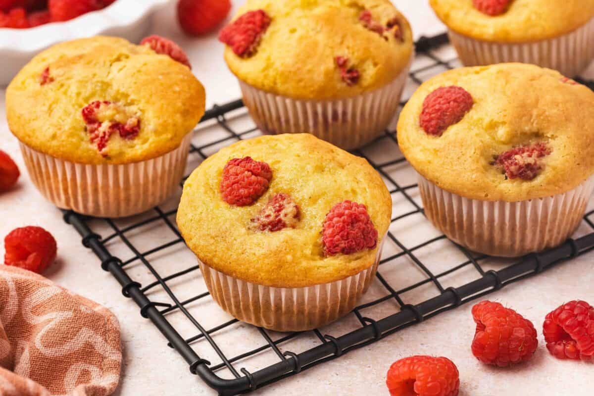 Five raspberry muffins in white paper liners rest on a black cooling rack, with fresh raspberries scattered around them on a light surface.
