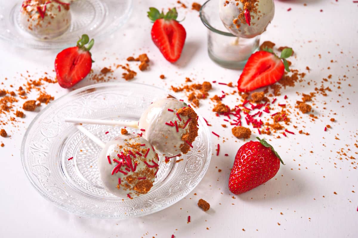 Two cake pops with white icing and sprinkles on glass plates, surrounded by fresh strawberries and cookie crumbs on a white surface.