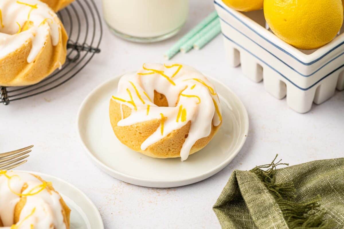 A lemon bundt cake with white icing and lemon zest sits on a small white plate, surrounded by fresh lemons, a glass of milk, and a green napkin.