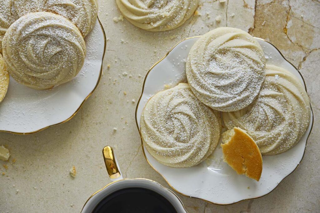 Plates of round, swirled shortbread cookies dusted with powdered sugar sit beside a cup of black coffee on a light-colored surface. One cookie is partially eaten.