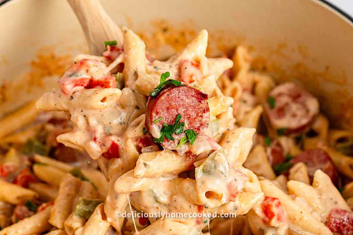 A wooden spoon lifts creamy penne pasta mixed with sausage slices, diced tomatoes, and herbs from a pot.