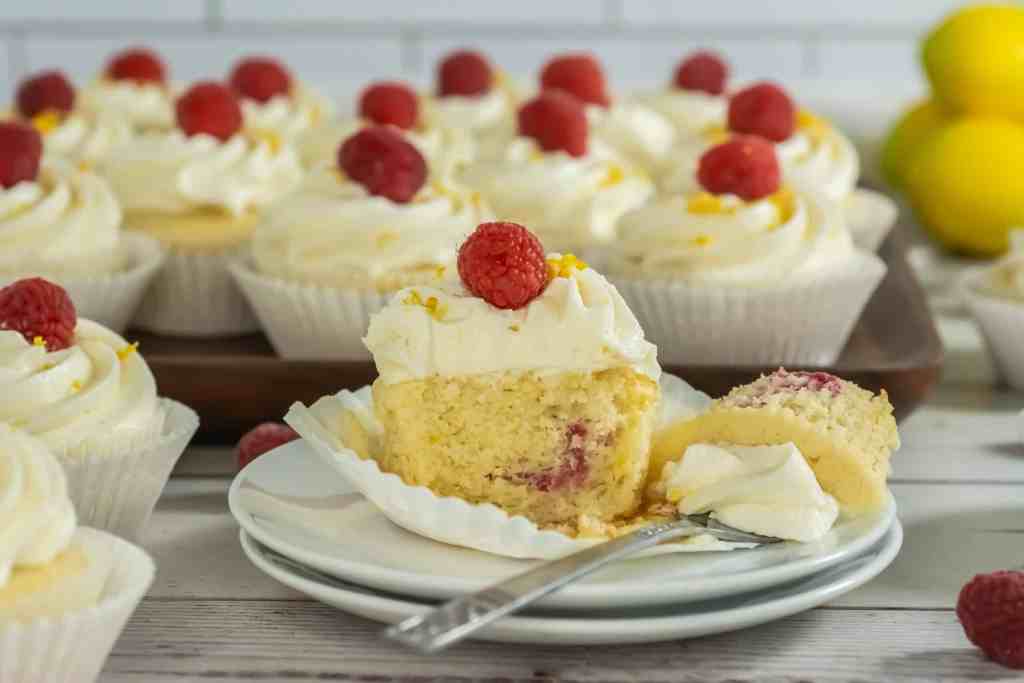 A plate with a partially eaten cupcake topped with frosting and a raspberry, surrounded by more similarly decorated cupcakes in the background.