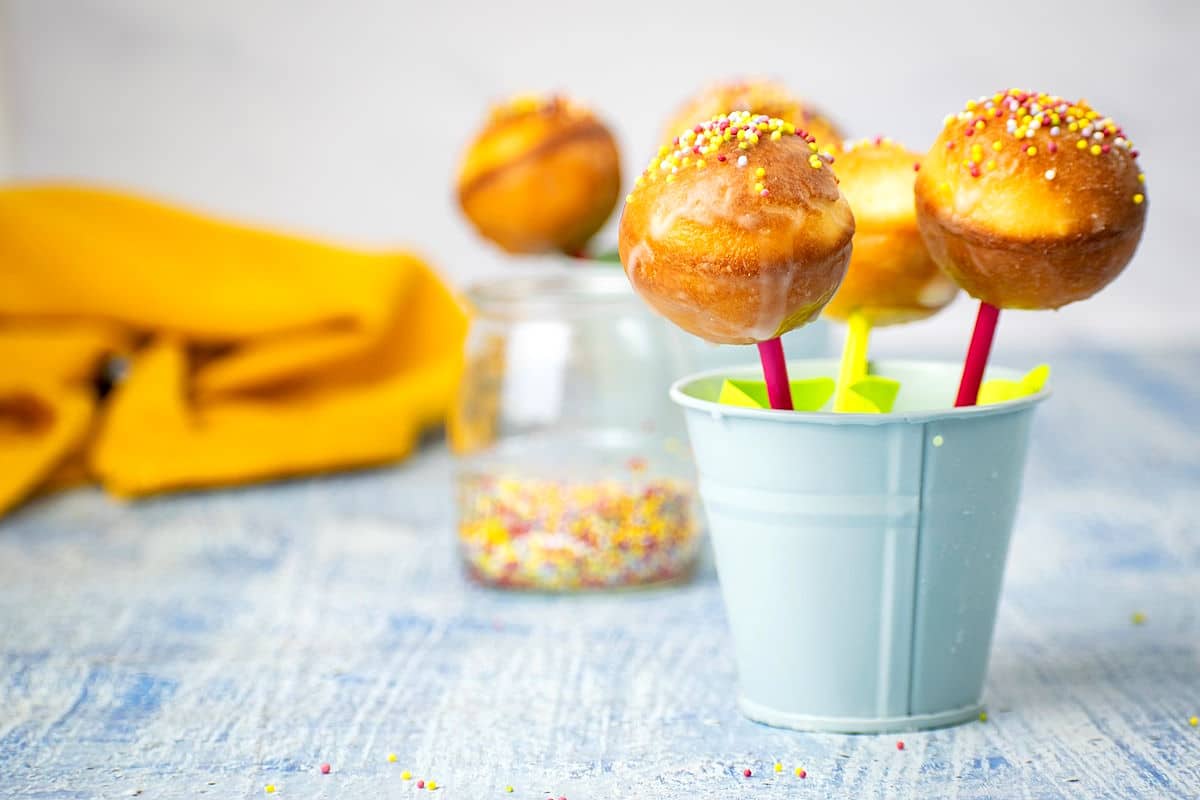Four cake pops with colorful sprinkles are displayed in a small blue bucket, with more cake pops and a jar of sprinkles in the background.