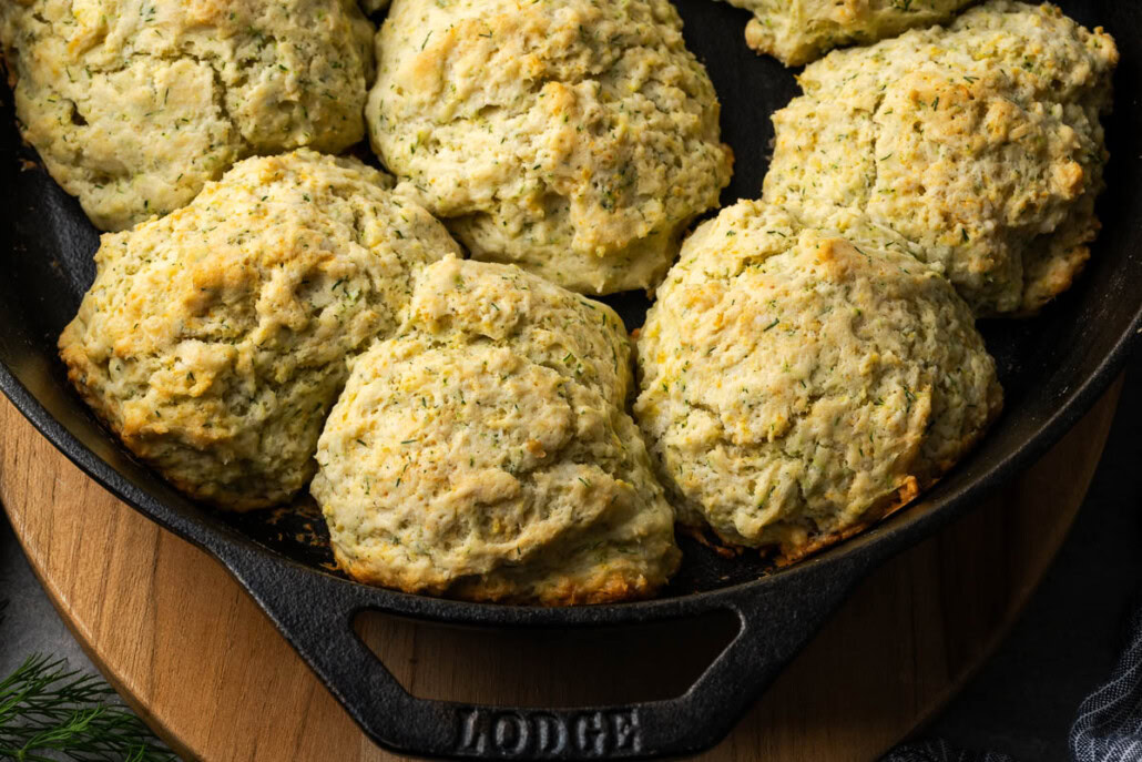 Seven golden-brown, drop-style biscuits are arranged in a cast iron skillet on a wooden board.
