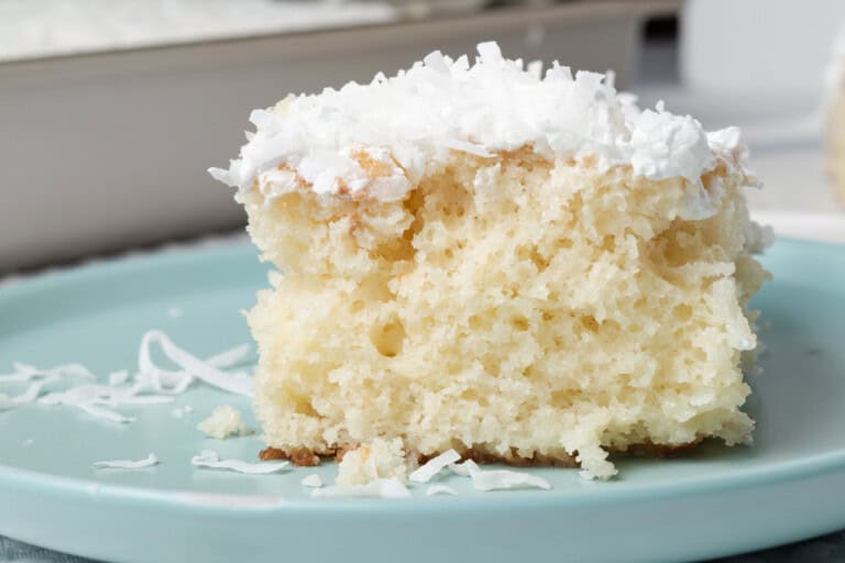 A slice of white cake with a fluffy texture, topped with white frosting and shredded coconut, sits on a light blue plate.
