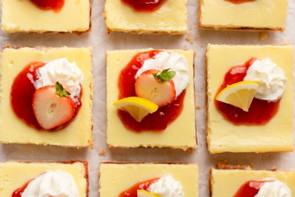 Lemon cheesecake bars topped with whipped cream, strawberry sauce, half a strawberry, and a lemon slice, arranged in neat rows on a light surface.