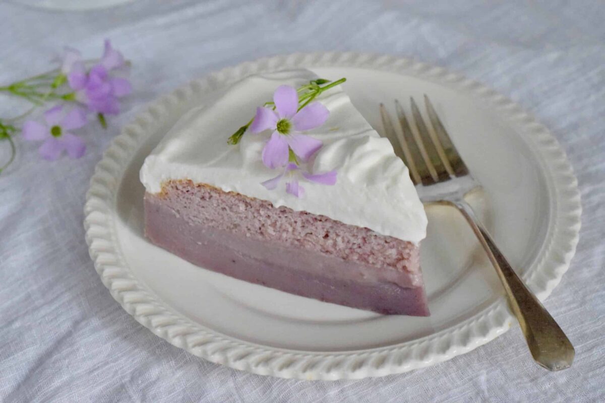 A slice of purple layered cake topped with white frosting and edible purple flowers on a white plate with a fork.