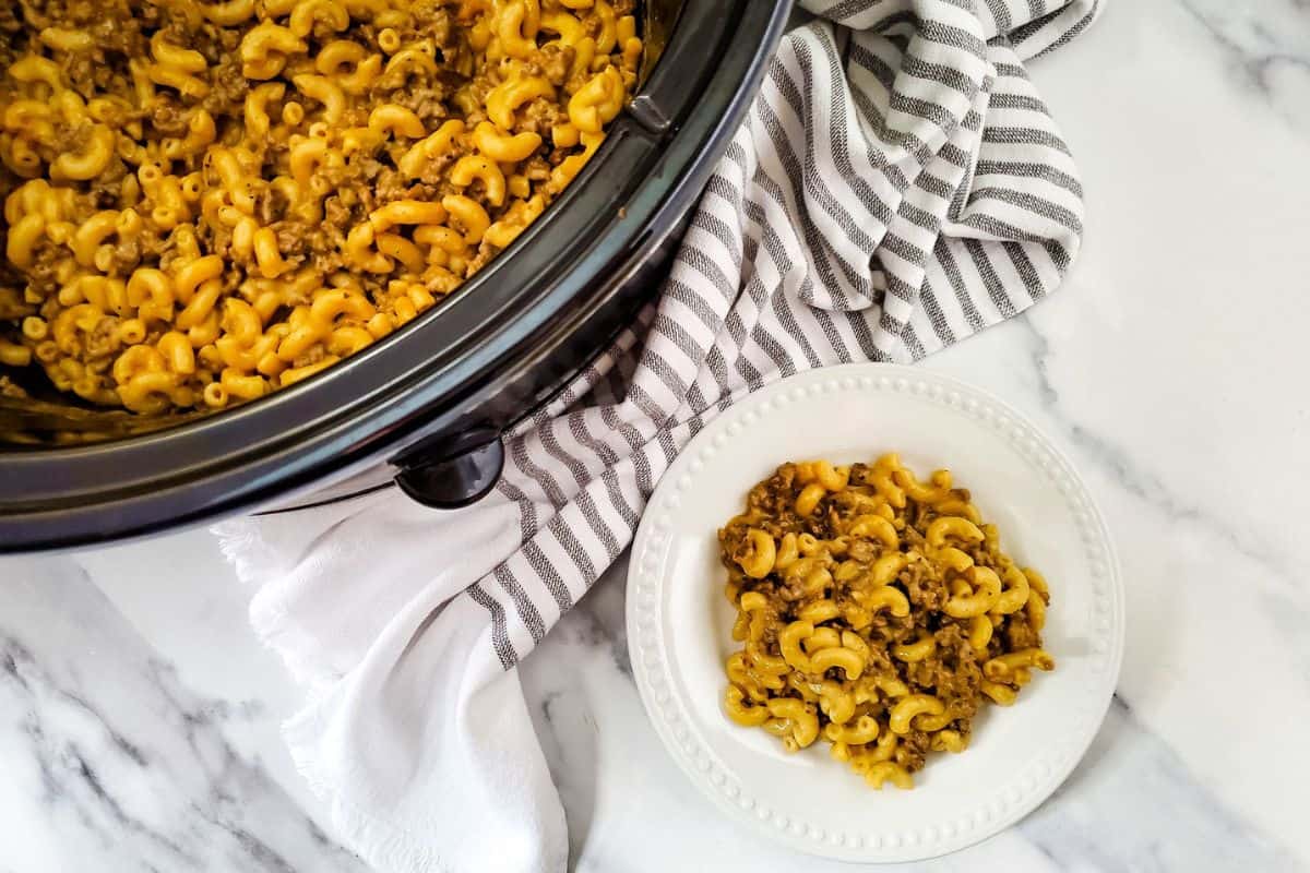 A slow cooker filled with macaroni and ground beef is next to a white plate with a serving of the same dish, set on a marble surface with a striped cloth.