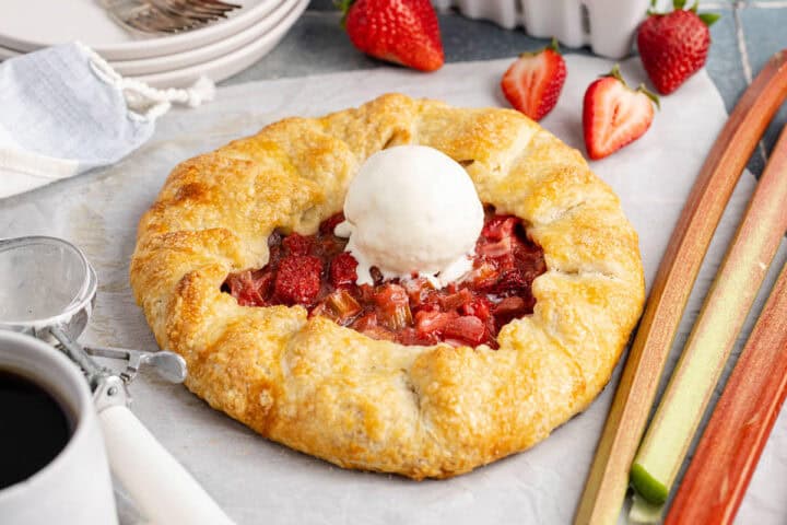 A rustic strawberry rhubarb galette topped with a scoop of vanilla ice cream sits on parchment paper, with fresh strawberries and rhubarb nearby.