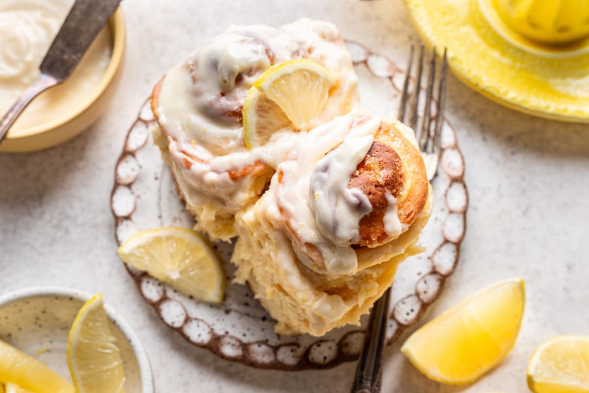 Two iced lemon rolls on a decorative plate, garnished with lemon slices. A fork, lemon wedges, and a bowl of icing are nearby on the table.