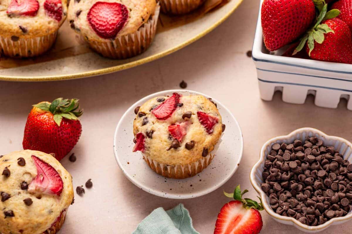 A plate with chocolate chip muffins topped with sliced strawberries, fresh strawberries, and a bowl of chocolate chips on a light-colored surface.