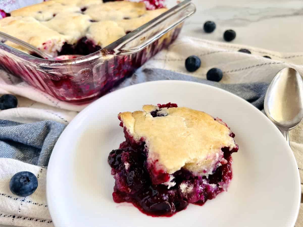 A serving of blueberry cobbler on a white plate sits next to a glass baking dish with more cobbler; scattered blueberries and a spoon are nearby.